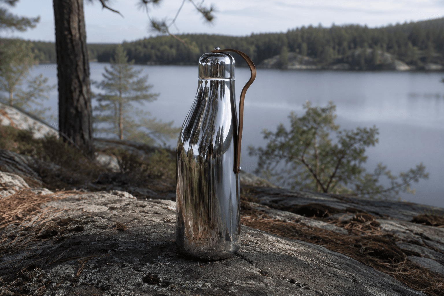 A polished silver flask with a brown leather strap on a lakeside rock